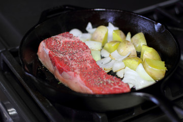 New York strip steak frying in a cast iron pan with potatoes and onions on the stove top.