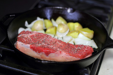 New York strip steak frying in a cast iron pan with potatoes and onions on the stove top.