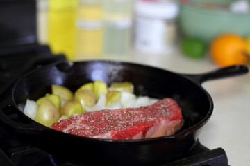 New York strip steak frying in a cast iron pan with potatoes and onions on the stove top.