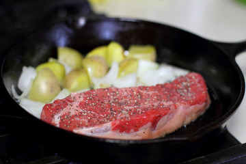 New York strip steak frying in a cast iron pan with potatoes and onions on the stove top.
