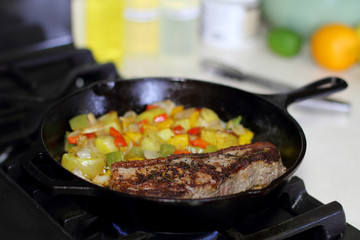 New York strip steak frying in a cast iron pan with potatoes,assorted bell peppers, and onions on the stove top.