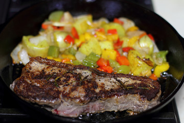 New York strip steak frying in a cast iron pan with potatoes,assorted bell peppers, and onions on the stove top.