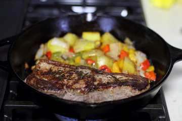 New York strip steak frying in a cast iron pan with potatoes,assorted bell peppers, and onions on the stove top.