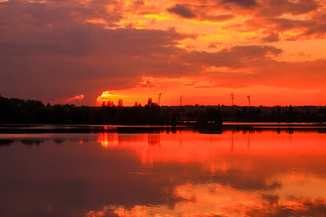 Beautiful dark sunset on the river with symmetry of colorful clouds cumulonimbus. Golden orange reflection of the sun's rays in water. Isolated tower on the skyline