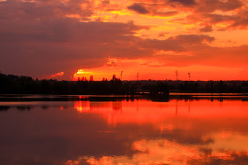 Fototapeta premium Beautiful dark sunset on the river with symmetry of colorful clouds cumulonimbus. Golden orange reflection of the sun's rays in water. Isolated tower on the skyline