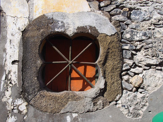 and ancient stone oblong shaped window with crossed bars with an orange shutter set in an old patched repaired grey wall with cracked cement