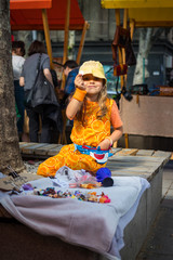 Little cute smiling girl shows her first money coin proud of experience of selling her old toys on local flea market. In dress and hat on a bench