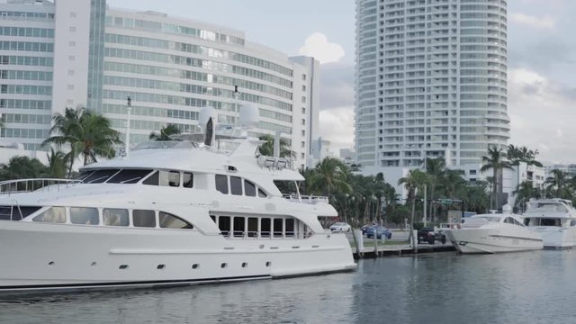 Luxury yachts parked in a Miami dock with a road with cars, palm trees and skyscrapers in the background under a cloudy sky in summer. 4K. Taken from a boat.