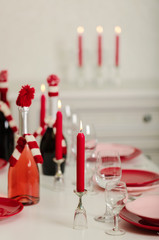 Merry Christmas and Happy New Year! Table setting - red and pink dishes, holiday knitted decor - Santa Claus knitted hats on the bottle with wine, red candles.