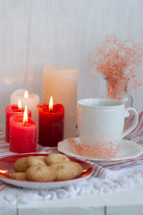 A hot cup of black tea with homemade cookies on a striped tablecloth, wax candles, a glass vase with decorative herbs on a white background.