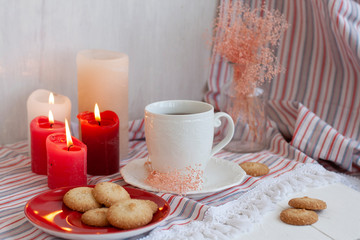 A hot cup of black tea with homemade cookies on a striped tablecloth, wax candles, a glass vase with decorative herbs on a white background.