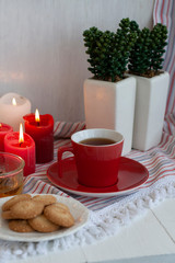 A cup of black tea with homemade cookies on a striped tablecloth, a bowl honey, a pair of decorative plants in a pot, wax candles on the white background.