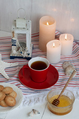 A cup of black tea with homemade cookies on a striped tablecloth, a bowl honey, a decorative lantern, a starfish, seashells; wax candles on the white background.