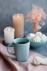 A hot cup of black tea with a bowl of airy meringues on a pink tablecloth, wax candles, a glass vase with decorative herbs on a gray background.
