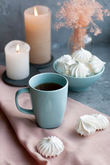A hot cup of black tea with a bowl of airy meringues on a pink tablecloth, wax candles, a glass vase with decorative herbs on a gray background.