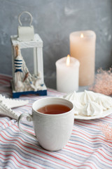A hot cup of black tea with a plate of airy meringues on a striped tablecloth, decorative starfish and lantern, wax candles, decorative herbs on a gray background.