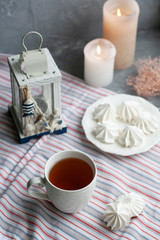 A hot cup of black tea with a plate of airy meringues on a striped tablecloth, decorative starfish and lantern, wax candles, decorative herbs on a gray background.