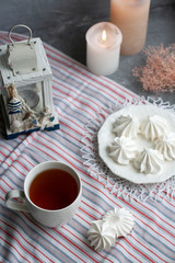 A hot cup of black tea with a plate of airy meringues on a striped tablecloth, decorative starfish and lantern, wax candles, decorative herbs on a gray background.