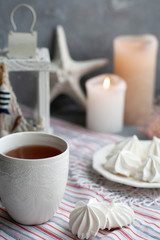 Obraz na płótnie Canvas A hot cup of black tea with a plate of airy meringues on a striped tablecloth, decorative starfish and lantern, wax candles, decorative herbs on a gray background.