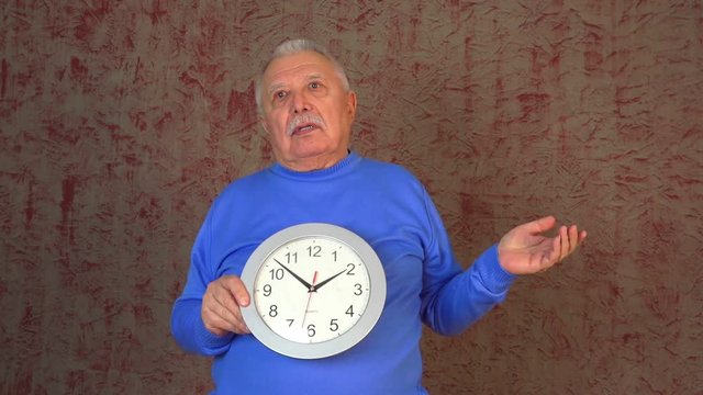 elderly man in blue sweatshirt with grey hair and moustache holds and resents wall clock closeup