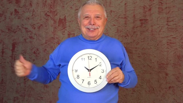 handsome old man with grey hair and moustache holds new silver clock surprises and shows cool sign thumbs up closeup