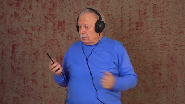interesting and joyful old man in blue sweatshirt searches music in smartphone turns on and dances close view