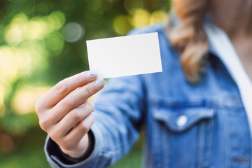 The girl holds in her hand a business card a mock up for the presentation of the design. Female hand holding a white business card with copy space on a green background of nature