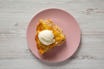 Slice of homemade apple pie with ice cream on a pink plate over white wooden surface, overhead view.