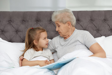 Cute girl and her grandmother reading book on bed at home