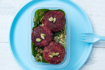 a portion of beetroot cutlets with pumpkin seeds in a plastic box on a plastic blue plate on a white table. have dishes plastic blue a fork