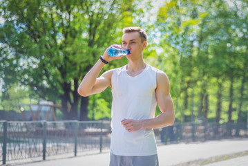 Sport man in white sportswear resting after outdoor workout, drinking water from bottle at park