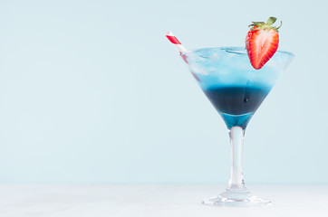 Cold tropical blue drink with ice, red straw, strawberry in misted wineglass in modern pastel green interior on white wooden table.