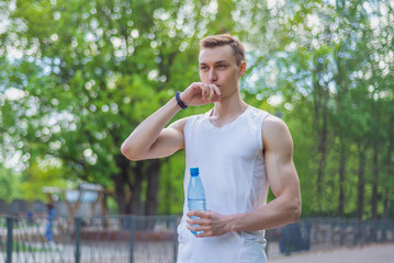 Sport man in white sportswear resting after outdoor workout, drinking water from bottle at park