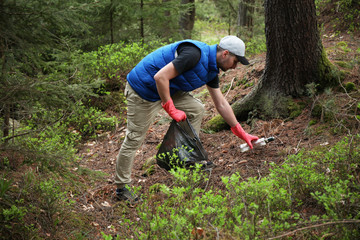 Naklejka premium Adult man volunteer in red gloves removes plastic trash in forest, caring for environment.