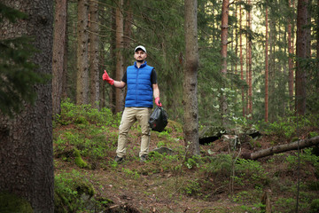 Adult man volunteer in red gloves removes plastic trash in forest, caring for environment.