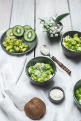 on a white, wooden table top, green tableware with fresh yoghurt and pieces of kiwi fruit sprinkled with coconut flakes, next to a tiny bouquet of field flowers and leaves in green and white color
