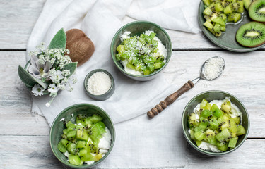 on a white, wooden table top, green tableware with fresh yoghurt and pieces of kiwi fruit sprinkled with coconut flakes, next to a tiny bouquet of field flowers and leaves in green and white color