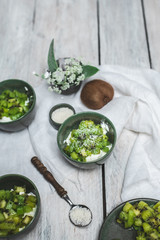 on a white, wooden table top, green tableware with fresh yoghurt and pieces of kiwi fruit sprinkled with coconut flakes, next to a tiny bouquet of field flowers and leaves in green and white color