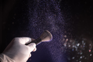 Makeup brush in hand with cosmetic powder on dark background with light and smoke. Powder splash on dark