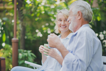 Closed up senior Caucasian couple talking with holding a cup of coffee on hand and felling happy in green garden. Lunch or tea time on picnic table in summer. Big family outdoor party concept.