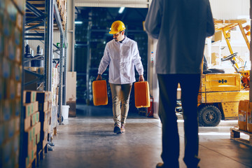 Young Caucasian employee dressed in white uniform and with protective helmet on head relocating oil canisters in warehouse while manager controlling him.