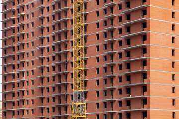 Close-up on a multi-storey residential building under construction from red brick with a part of a yellow crane. The walls of the house in the process of work and architecture.