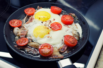 scrambled eggs cooking in a frying pan, cooking on a ceramic stove, fried eggs with bacon and tomato, 45 view closeup