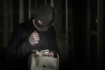 Robber in a mask and with a flashlight examines the contents of the women's bag in a dark room