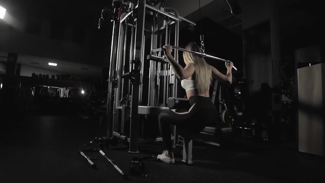 Fitness woman training with weight-lifting training machine in slow motion in dark gym. Blonde girl doing exercise on shoulders in block simulator