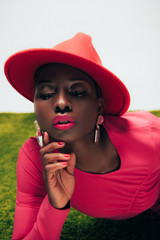 beautiful stylish african american woman in pink dress and hat posing on green grass