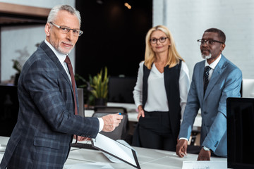 selective focus of cheerful businessman in glasses holding clipboard near multicultural partners in office