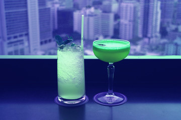 Two different type of cocktails in vibrant green color on the rooftop bar's table with blurry skyscrapers view in background