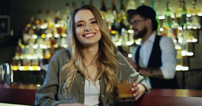Portrait shot of the beautiful blond Caucasian woman smiling to the camera while sitting in the bar with alcohol drink, cocktail or cider.