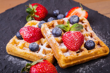 Homemade Traditional belgian waffles with fresh fruit, berries and sugar powder on black plate. Flat lay, top view, copy space.